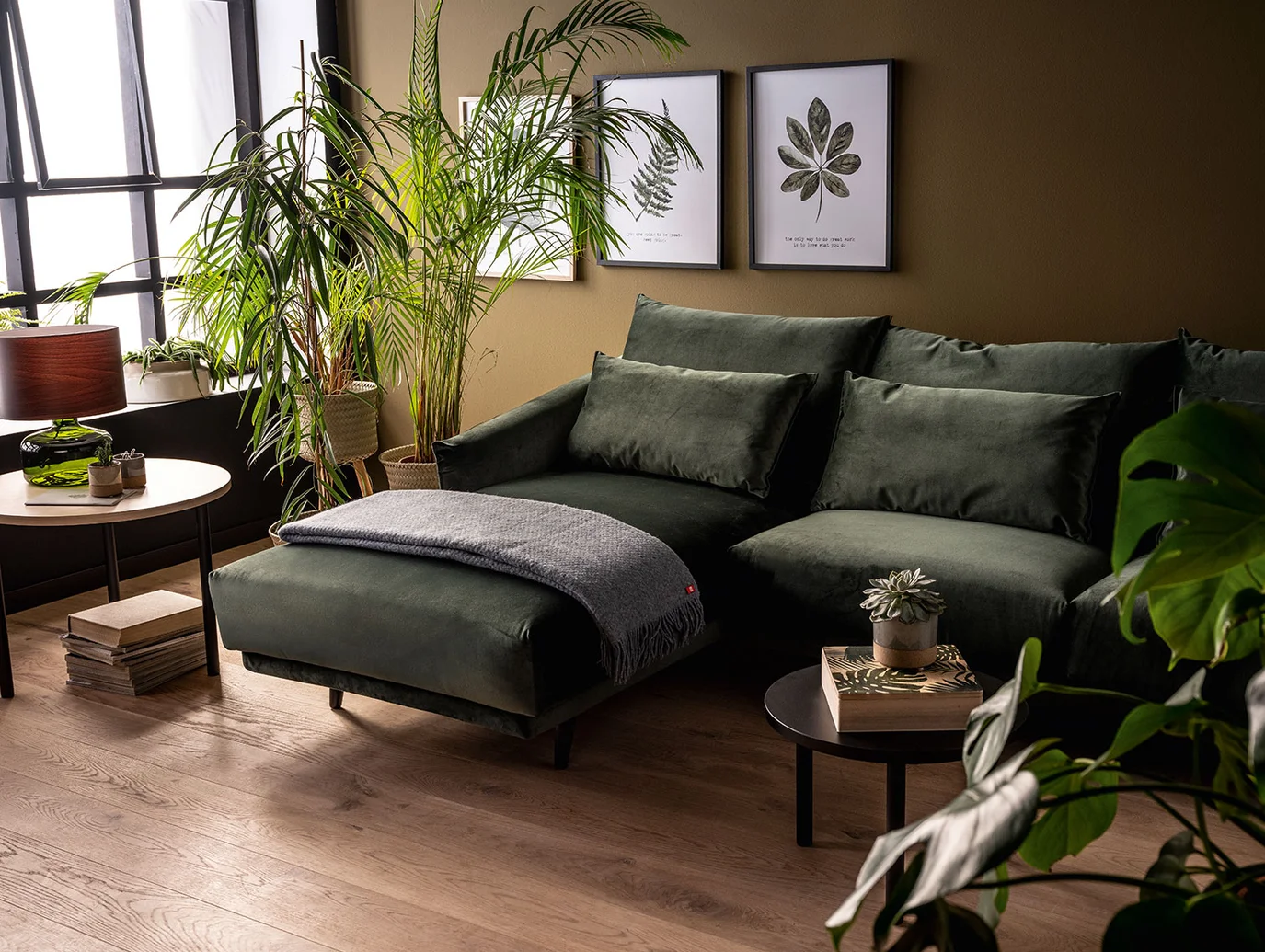 Zielony narożnik w salonie z licznymi roślinami doniczkowymi i botanicznymi grafikami na ścianie.